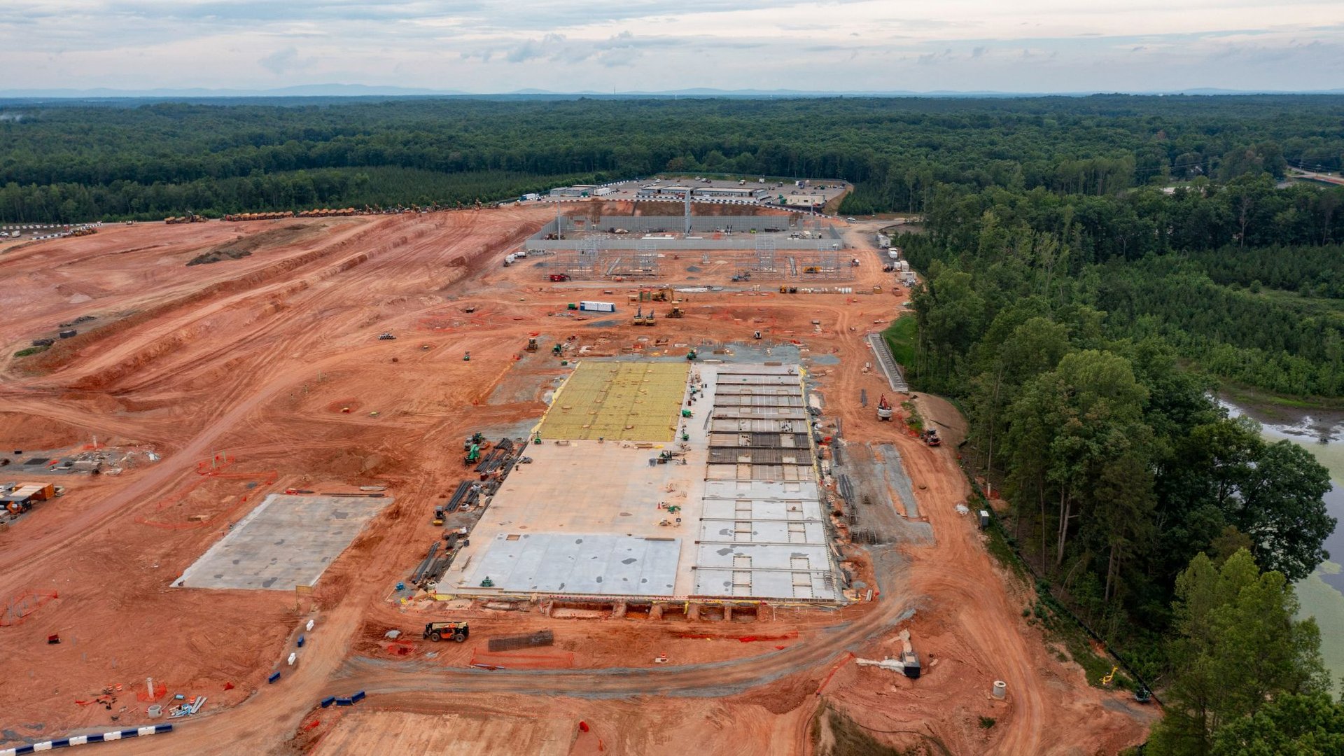 Aerial view of a large construction site with poured foundations and red dirt bordered by dense forest on both sides
