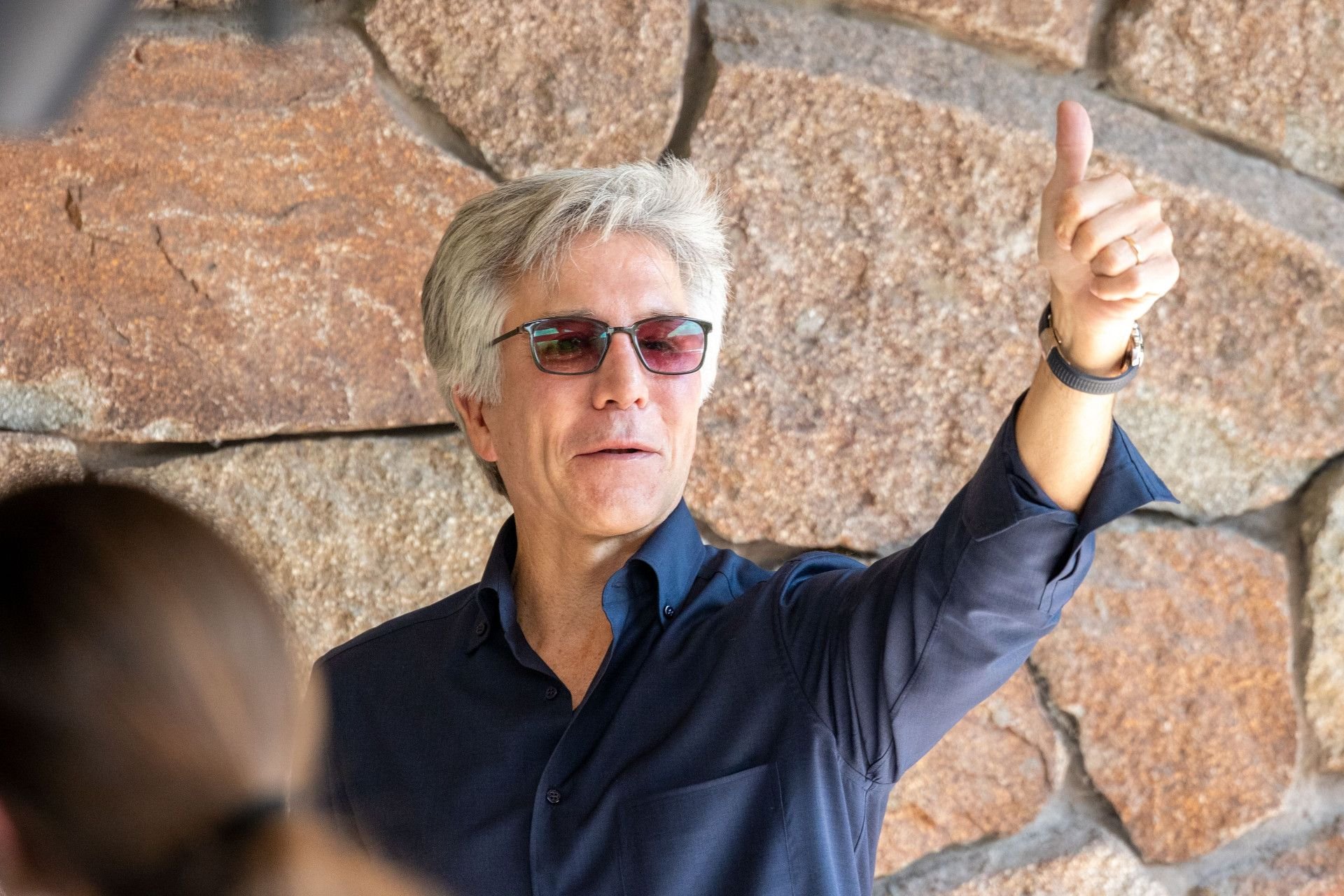 Man with gray hair and sunglasses giving a thumbs up gesture against a stone wall wearing a dark blue shirt looking confident and positive in an outdoor setting