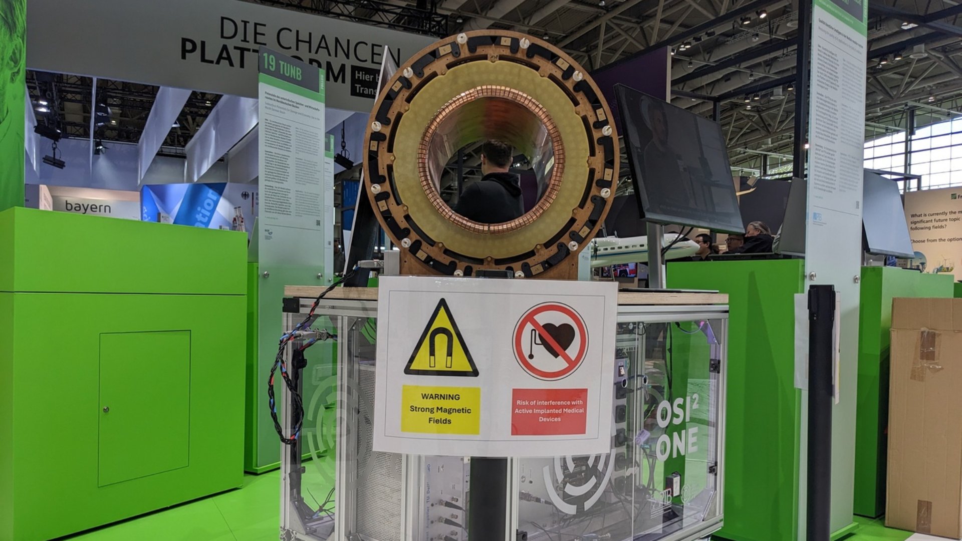 Exhibit at a science fair featuring a large donut shaped magnetic coil bright green display panels and warning signs in the foreground
