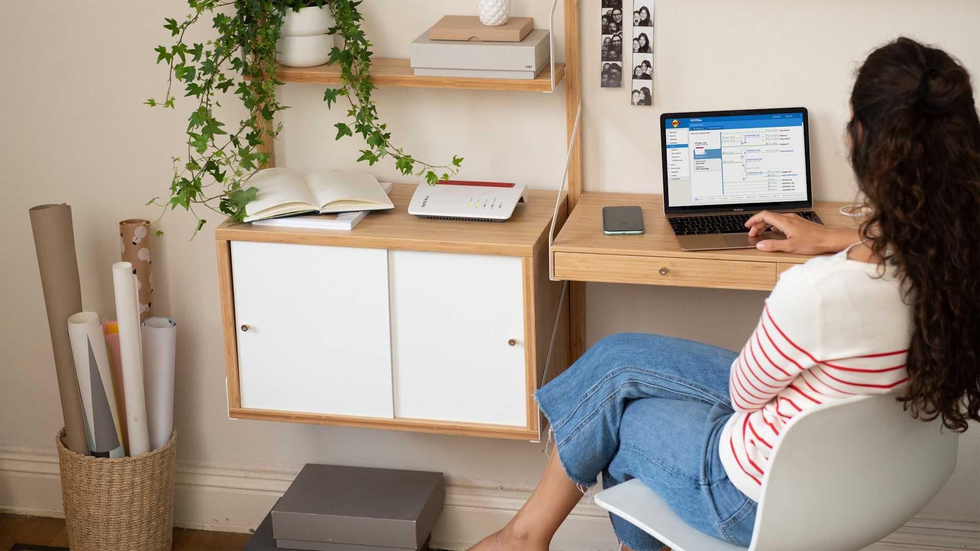 Person with long curly hair striped shirt and jeans sits at a wooden desk using a laptop in a bright home office with a potted plant and photo strip on the wall shelf