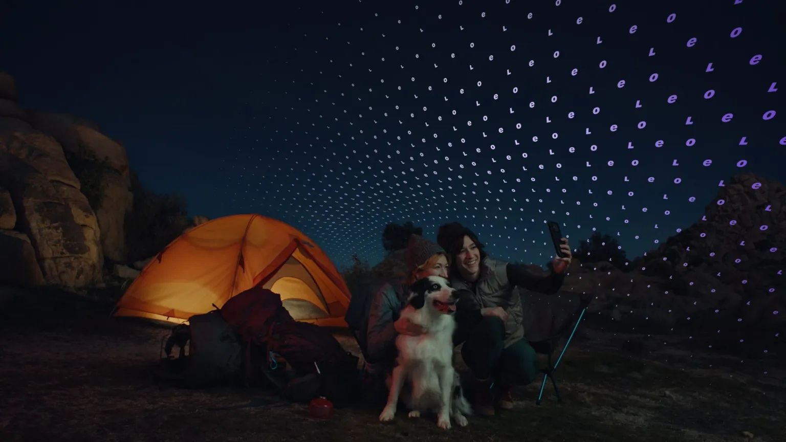 Two people sit by an orange tent at night posing for a selfie with a dog under a starry LED projection
