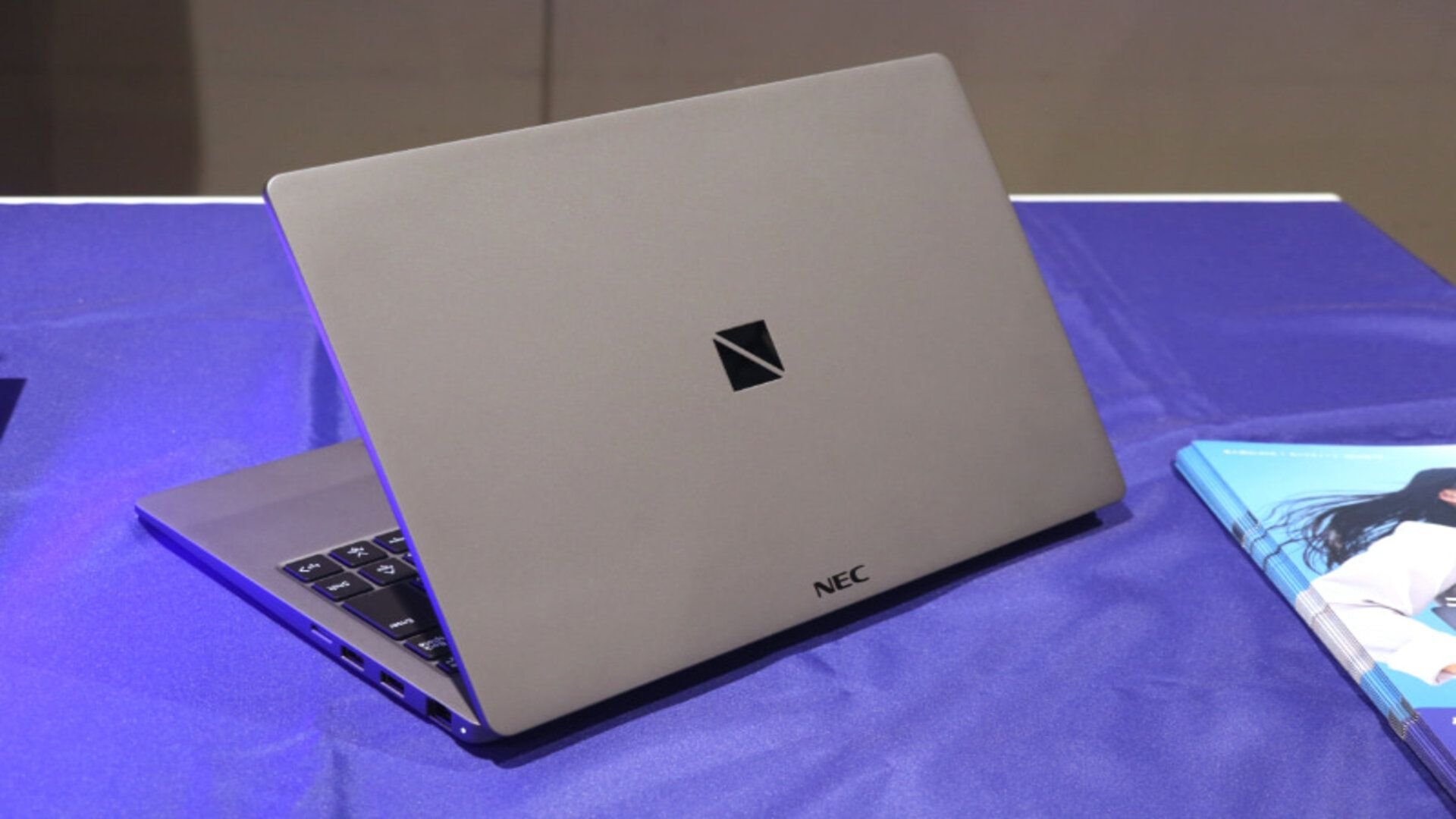 Slim silver NEC laptop open on a blue table keyboard visible along the left edge NE C logo on the back